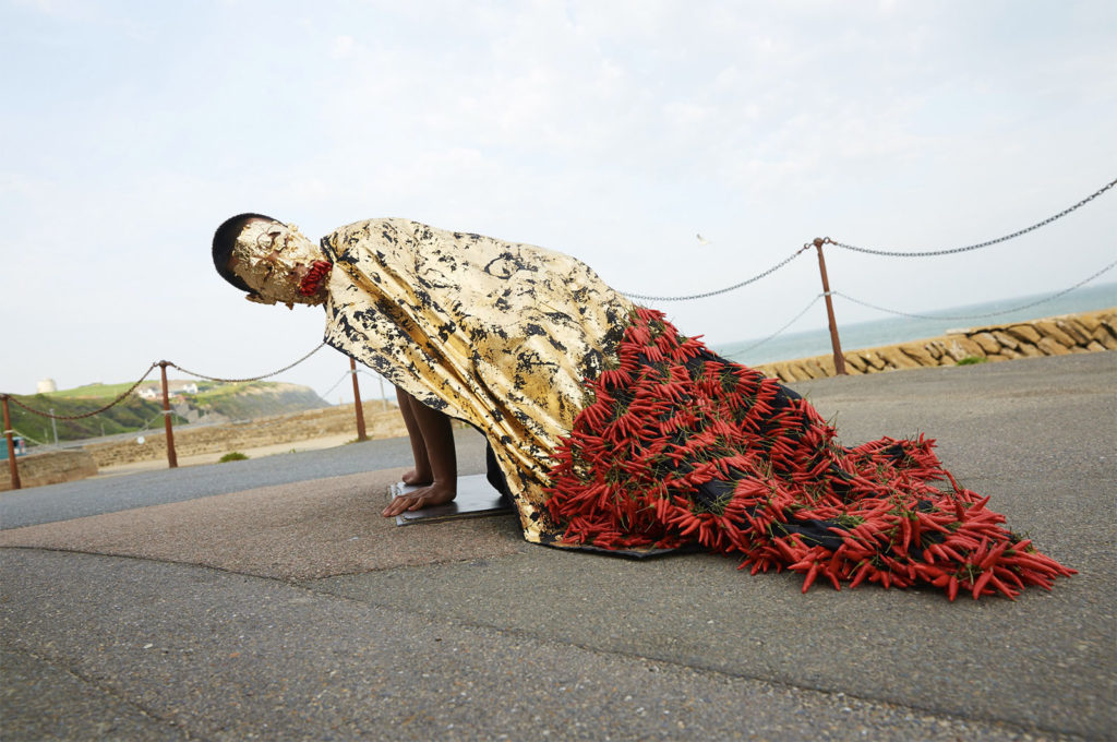 The artist, a twenty-something singaporean man, is on his hands and knees by the sea. He has closlely cropped black hair. His face is covered in gold leaf which is peeling. He wears a gold cloak which trails on the floor with many red chillis stitched into it. He also holds red chillis in his mouth.