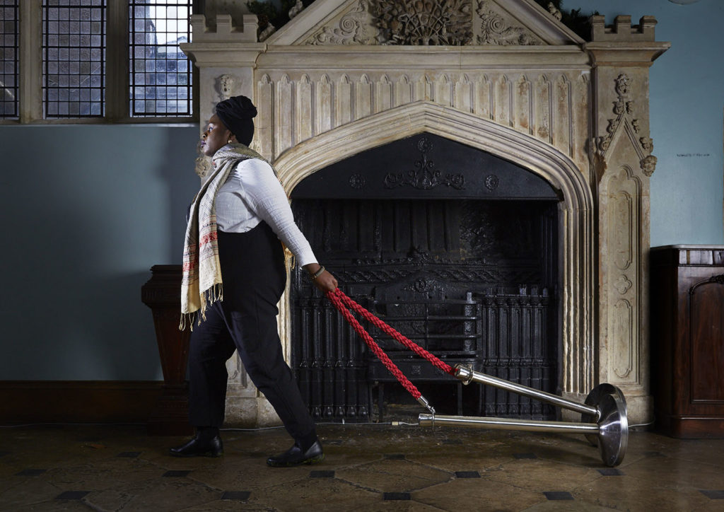 Muneera, a person with dark skin, walks through the corridor of a grand old english building. They pull the kind of cordon you might find at the theatre or cinema along the floor behind them. They appear determined and purposeful.