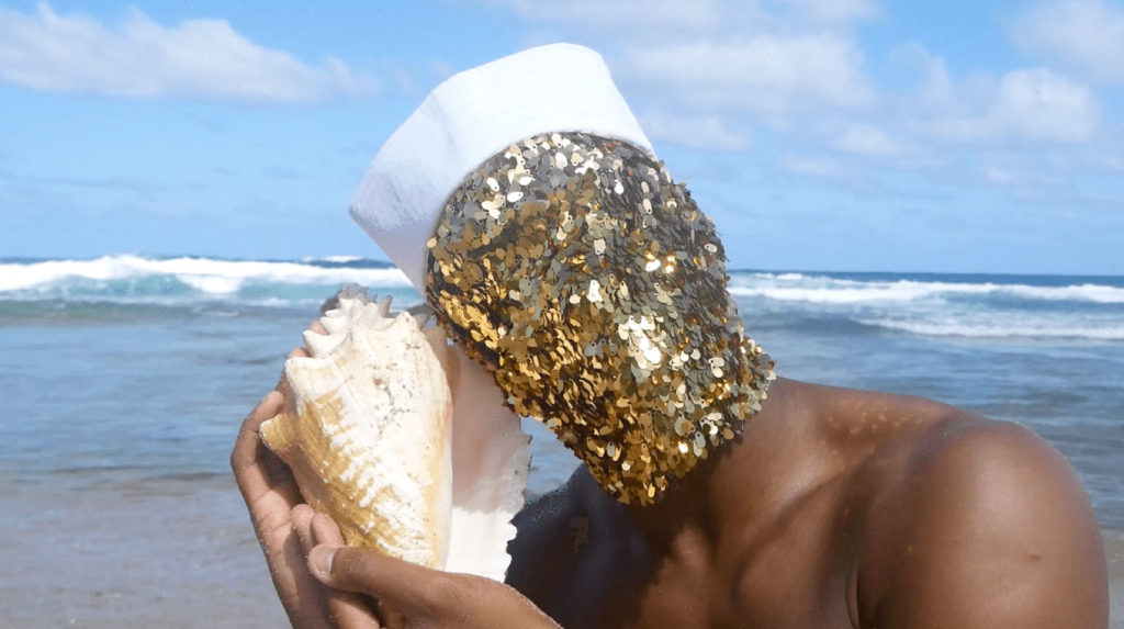 Ada, a black nonbinary person, stands at the sea shore in the carribbean. They wear a white sailors hat, and a mask of gold which reflects onto their skin. They hold a conch shell to their ear, listening to it.