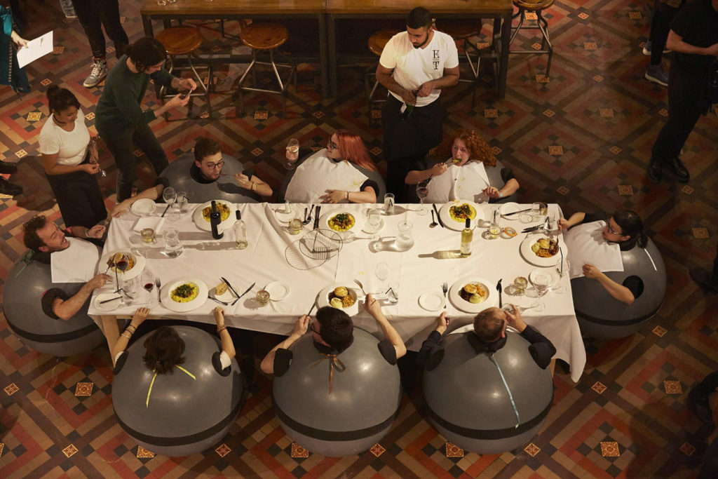 Audience are sat around a dinner table. They are sat close to the floor, inside spherical structures whilst museum staff deliver their food.