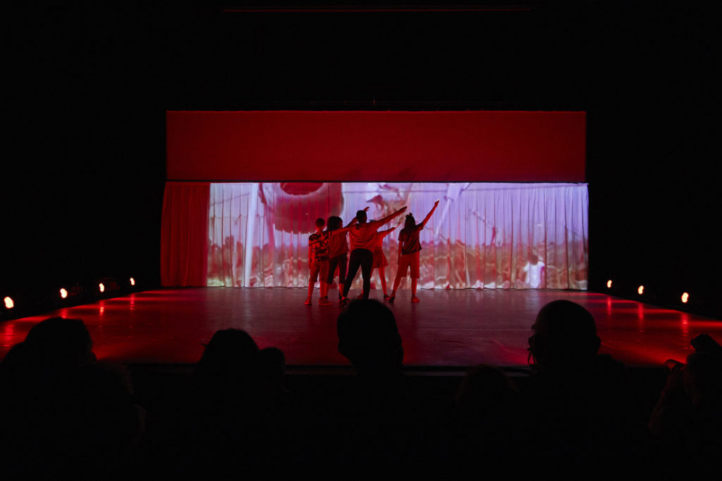 A group of 6 young people stand on stage in a dark theatre space. They are lit with red light, and face a projection across the rear of the stage.