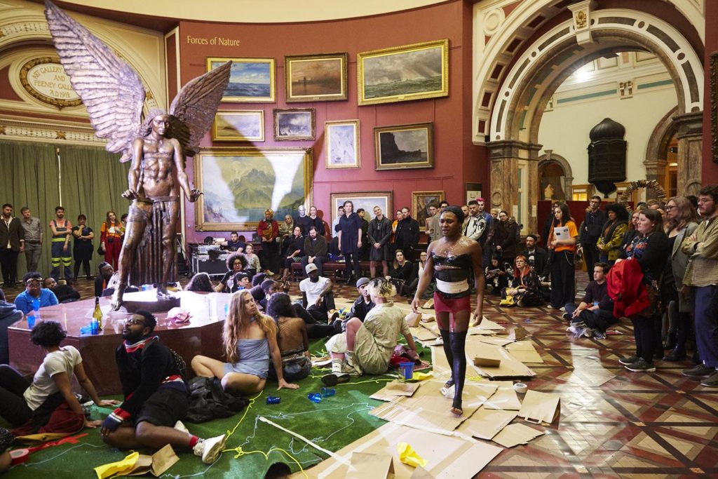 The artist, a black trans femme, circles a statue of an angel inside a museum. Keijaun wears black leggings and corset with a red girdle and black masking tape across her chest. The statue is dressed similarly. The floor is covered in cardboard and the audience surround the performance.