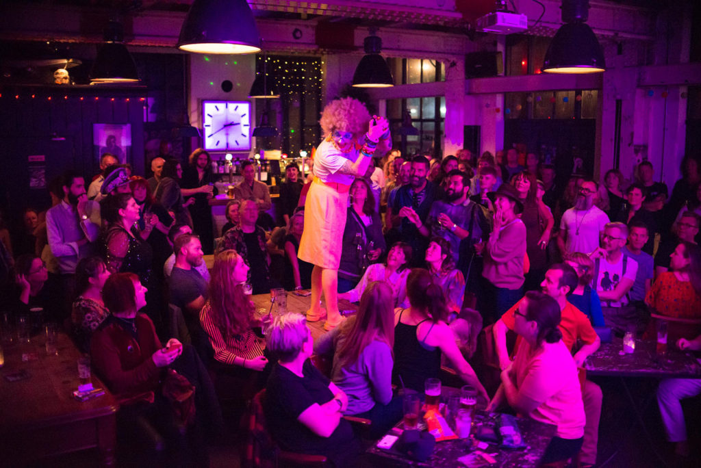 .Ginny Lemon, a drag performer with a bright yellow wig and skirt and clownish makeup stands on a table in a bar. They are lit by a strong purple light and are surrounded by audience.