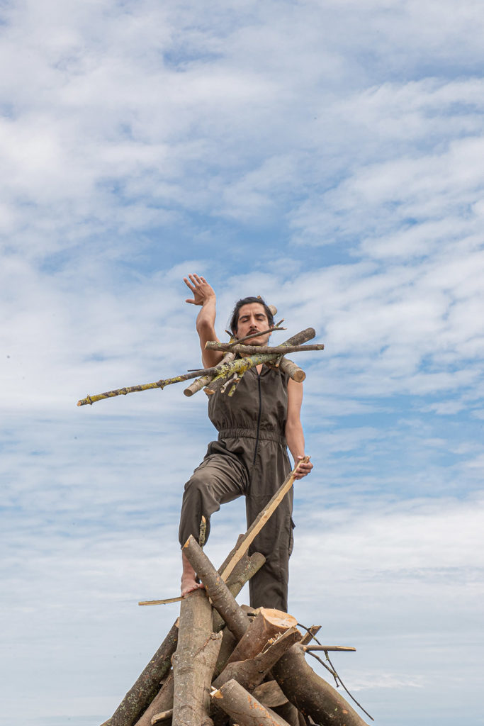 Emilio, a person with brown skin, black hair and a moustache stands on top of a pile of branches beneath a big blue sky. There are more branches balanced around Emilio's neck and they extend their arm towards the sky.