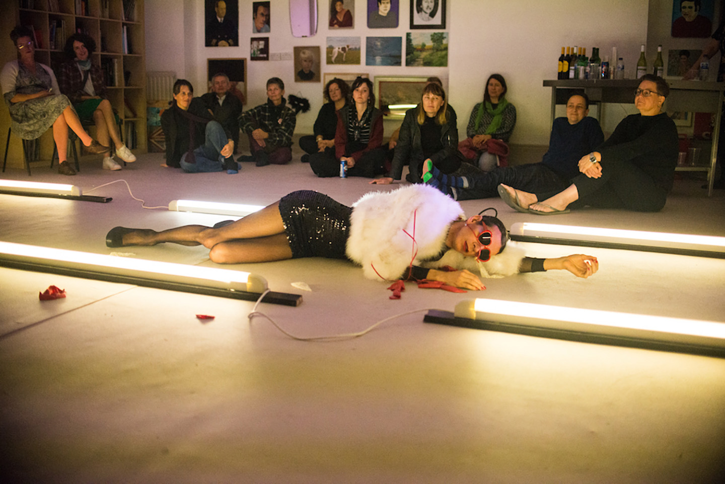  Nando, a femme presenting person, lies on the floor between strip lights. They wear a fluffy white coat, a black sparkly dress and red heart shaped sunglasses. They are surrounded by burst red balloons. An audience gazes at their prone form.