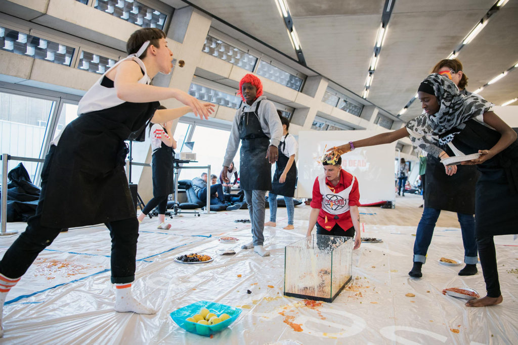 A transgenerational group of children and adult artists are working with food inside a museum gallery. One child wears a hijab, another wears a red wig. One of the adults is spitting food. There is tarp on the floor which protects it from the mess.