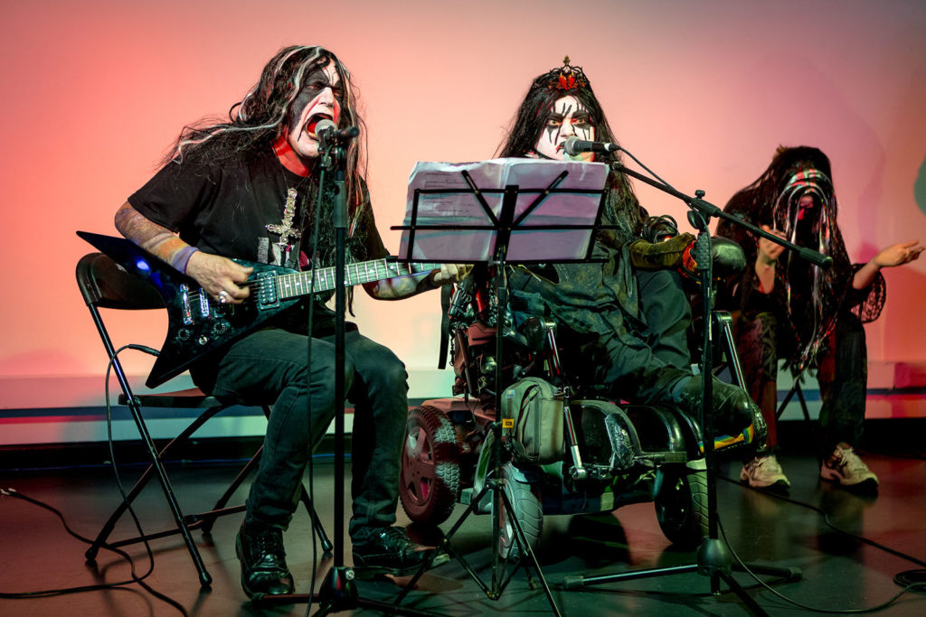 Katherine and Aaron, the Disabled Avant Garde, are dressed all in black, like members of the heavy metal band kiss. They also wear black and white wigs, and have black and white makeup on their faces. Katherine is in her motorised wheelchair. Aaron sits next to her and plays guitar. Both are singing into microphones. A third person is in the rear of the shot, also wearing a wig, and arms raised in worship.