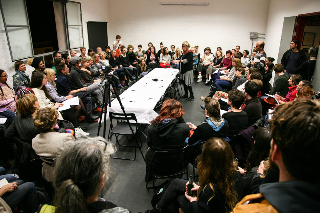 Lois Weaver, a white woman, stands by a long table covered in paper, and addresses a crowded room of participants (mostly women).