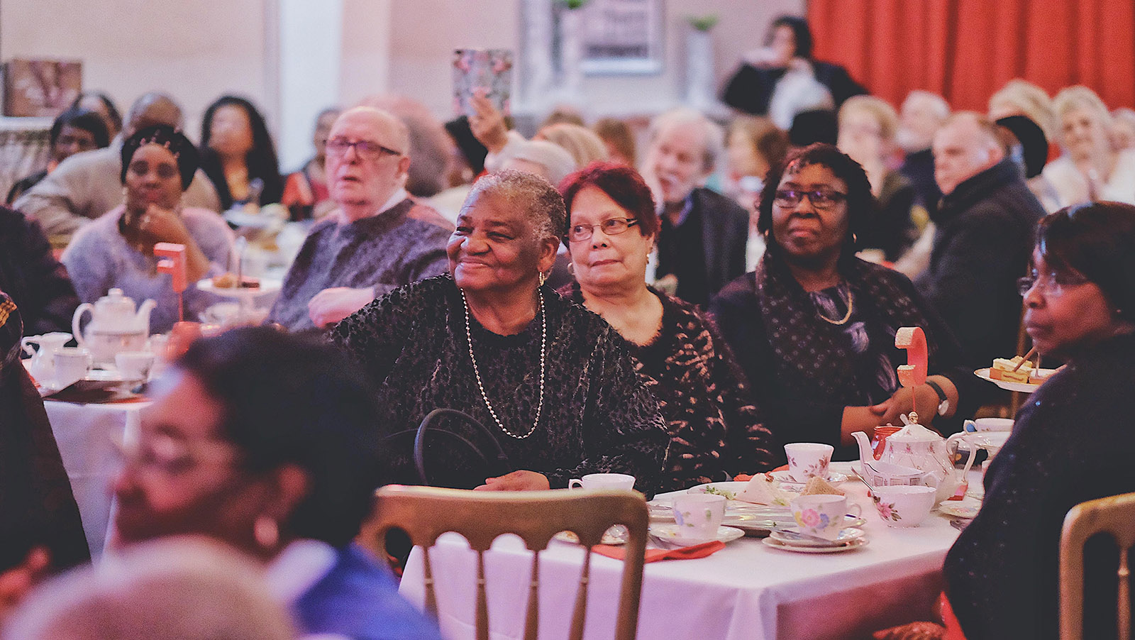 Audience members sit at tables with white tablecloths, fancy teapots and tea cups enjoying the stage show at the Posh Club. 