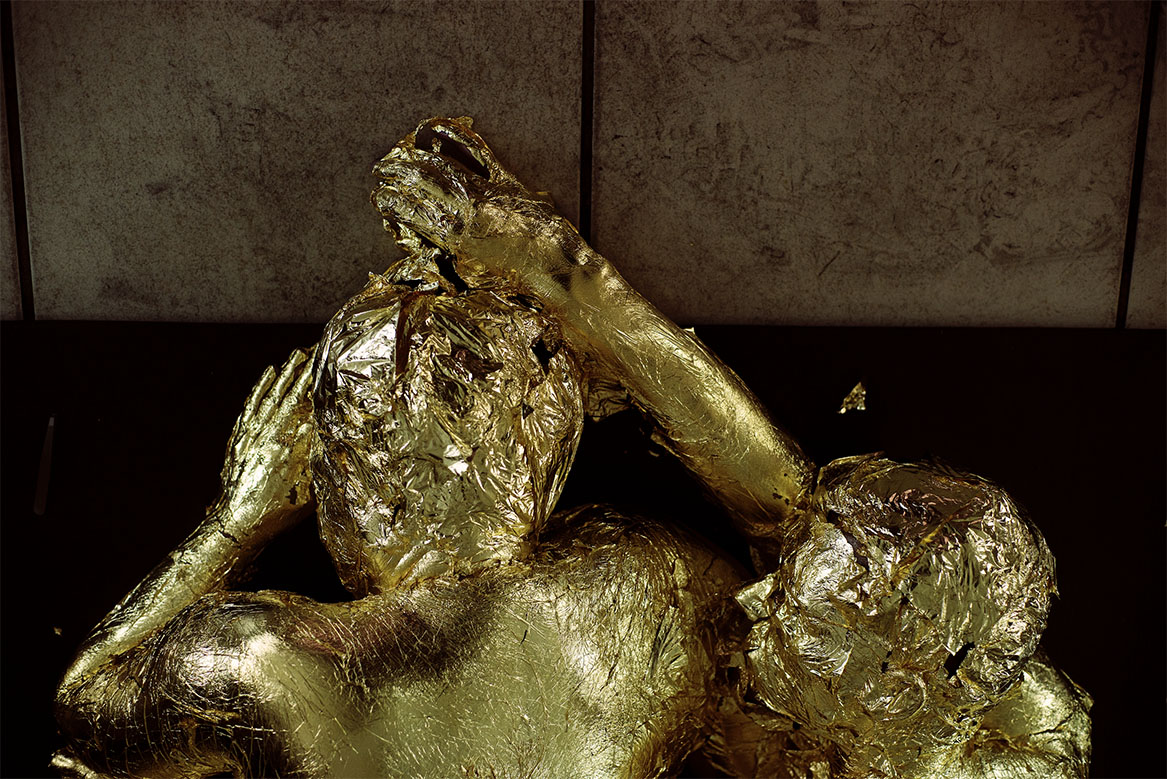 Two artists lie on a mat next to a tiled floor. The floor is covered in coal dust. They appear to have collapsed here, with one artist holding the other from behind. They are both completely covered in gold leaf.