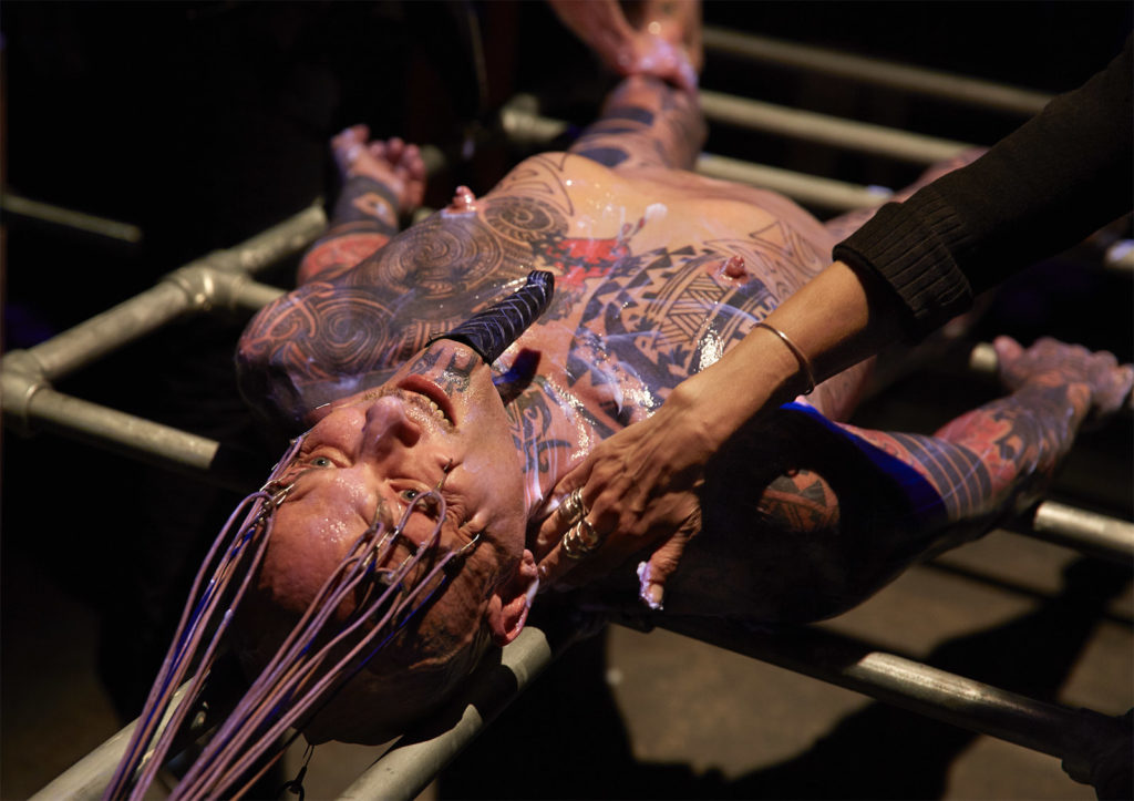 The artist, a middle aged white man, lies nude on a metal frame. He has many tattoos on his face, neck, and body. He wears a long false goatee, like those found in images of pharoahs. He is covered in a thick liquid, like oil or lube, and two people are rubbing it over his skin. There are hooks in his cheeks and eyebrows attached to wires which pull his face into a grimace.