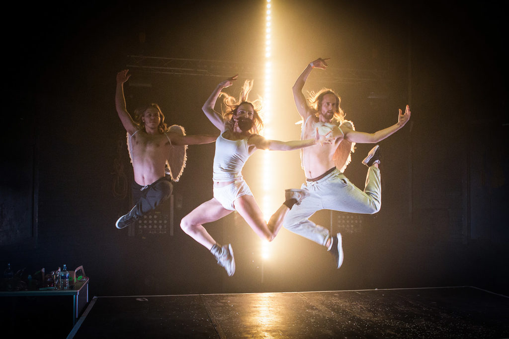 three people are jumping in unison on a dark stage with their arms raised above their heads. The people on the left and right are shirtless and wear white angel wings. The person in the centre wears a white tank top and briefs and a false beard. There is a bright strip of light running vertically behind them, lighting the stage