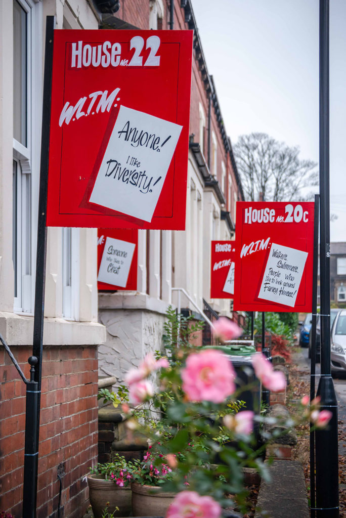 A row of red brick terraced houses in Leeds. There are red signs outside each advertising who the residents would like to meet. The sign in the foreground reads: "WLTM: Anyone! I like Diversity.