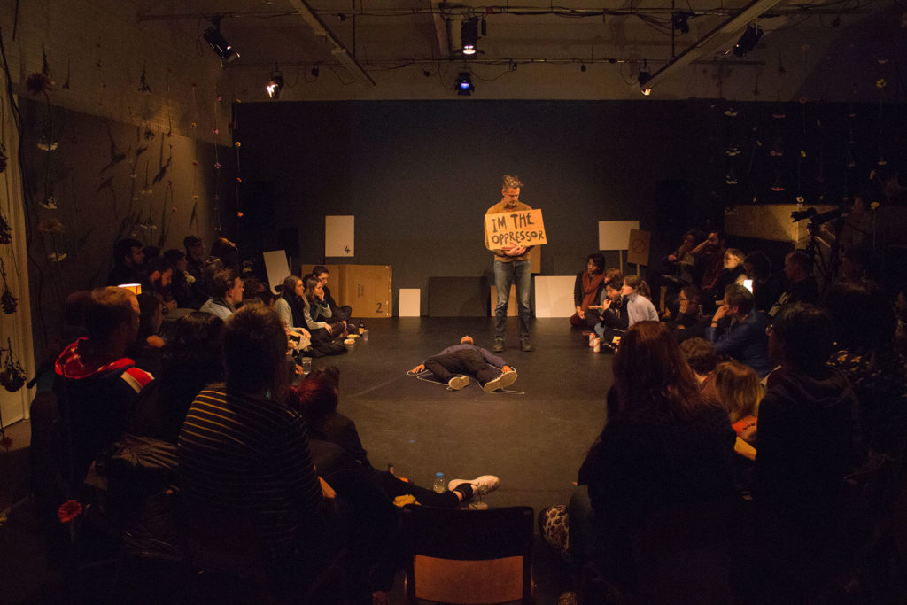 A dim lit performance space with audiences sitting on each side. A black person is lying in the center with a white outline drawn around their body. Standing beside them, a white person holding a cardboard cutout which says 'I'm the Opressor'