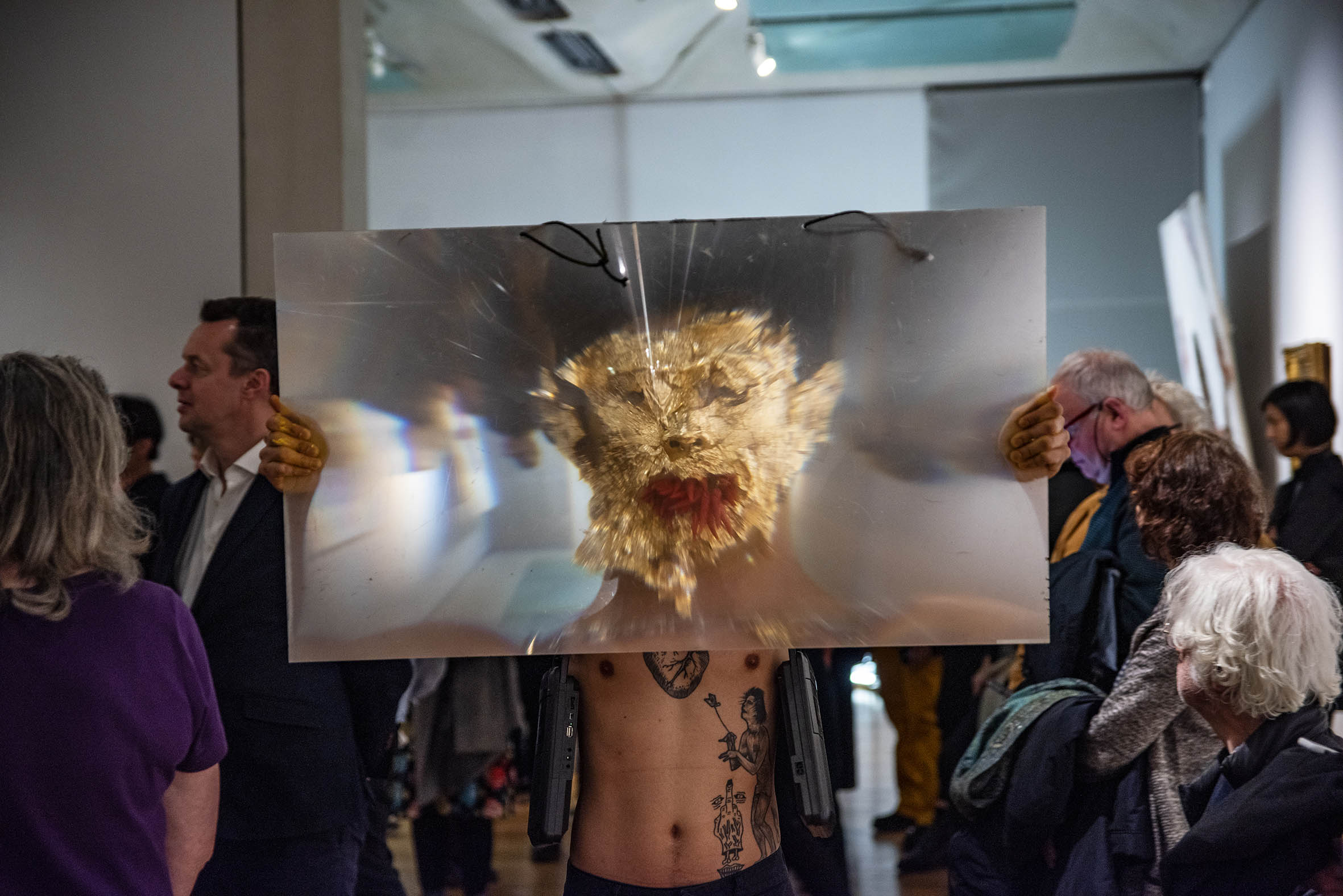 Nick, an asian person, is standing in a crowded gallery space, their body is covered in tattoos, two tape recorders dangle on their shoulders. They are holding a piece of magnifying plastic accentuating their face covered in golden foil and their mouth full of red chillies.