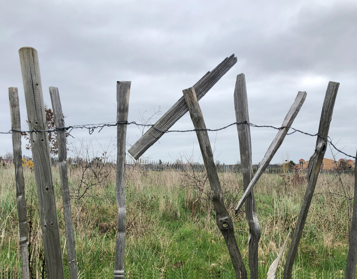 A broken fence in a field of green long grass. The grey pieces of wood are held together by a cord of barbed wire. The weather is cloudy and grey, in the distance there sits an orange building.
