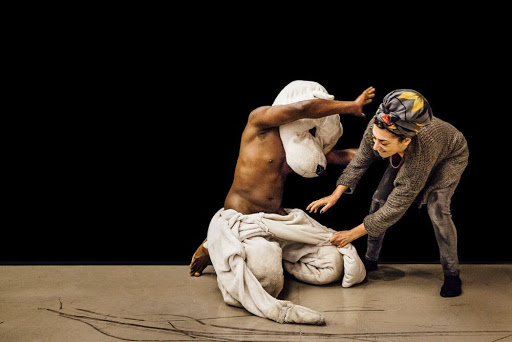 A black, male performer kneels on the floor wearing a white furry bear suit up to his waist, and a large white furry bear head obscuring his face. His chest is bare. A woman stands to his left, and tries to remove the rest of his costume.