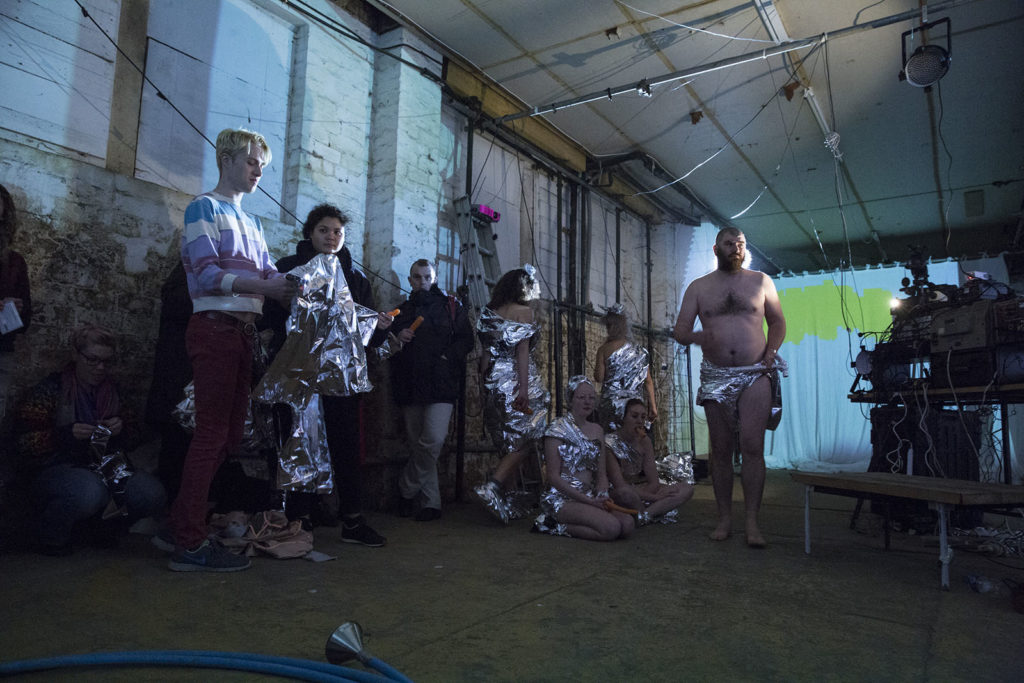 Daniel is stood in a disused warehouse space wearing nothing but a silver safety blanket as pants. He has a bald head and a beard. A collection of other performances are knelt and stood nearby, also wearing silver safety blankets. Some are holding carrots, one has a carrot in their mouth. There audience members are leant against a wall, holding safety blankets and carrots. A yellow splurge is projected against the back wall. 