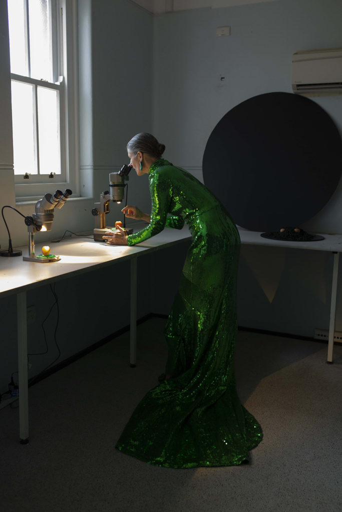 A woman with white skin in a long green sequined gown stands at a bench and peers down a microscope.