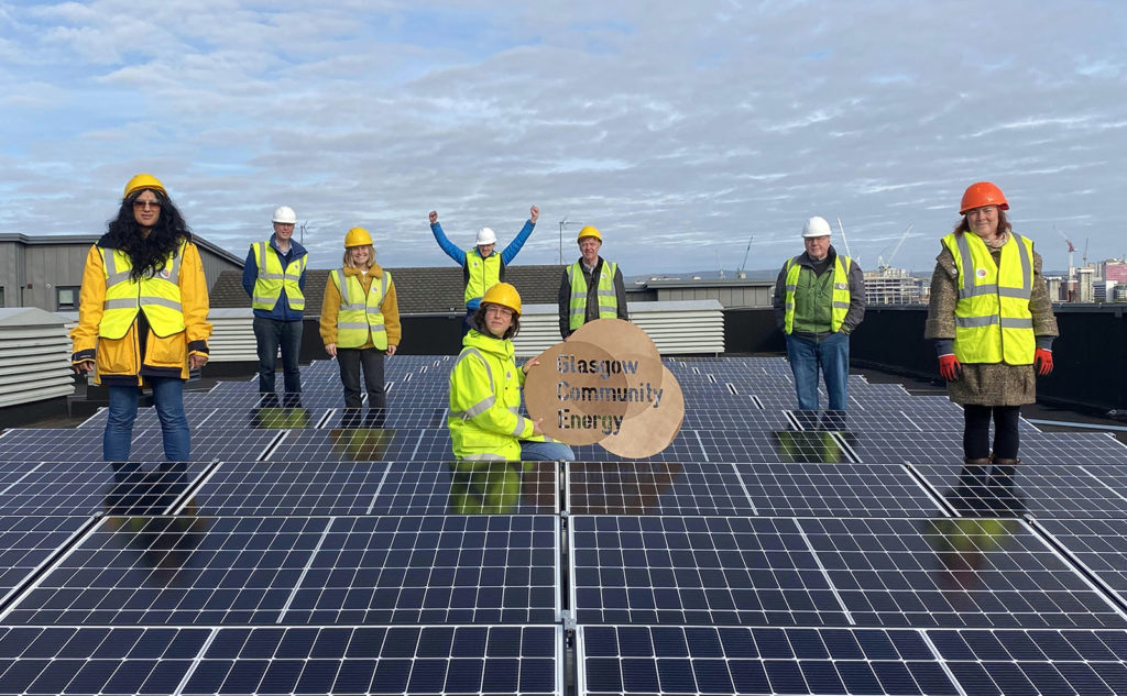 8 people in high vis and hard hats stand proudly in between a series of solar panels on a rooftop. Behind them is the sky and city skyline. Ellie holds up a sign that reads Glasgow Community Energy. 