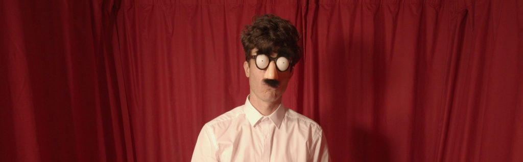 Tim stands in front of a red curtain with only his head and shoulders visible. He is wearing a fake nose and moustache. His eyes are covered with glasses that hold fake blank eyes. His lips are contracted.