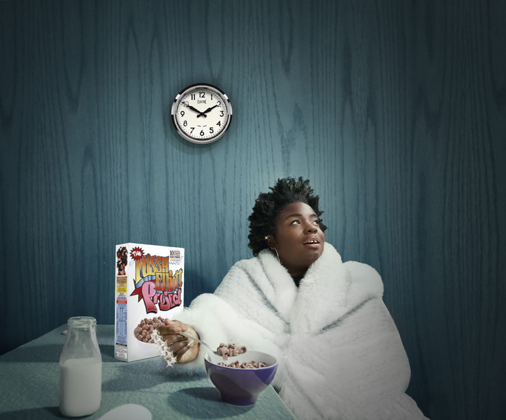 A dark skinned Black Woman with a short afro sits against a dark blue background at a dark blue table eating brown cereal from a purple bowl. She wears a large white fur coat, with a sparkling knuckle duster, a diamond helix piercing and gold hoop earrings. She is looking to the right with a slight smile, and her make up reflects early 90s Hip Hop Trends. On the table in front of her is a glass bottle of milk, a small milk puddle and a colourful cereal box that says 'The Missy Elliott Project'