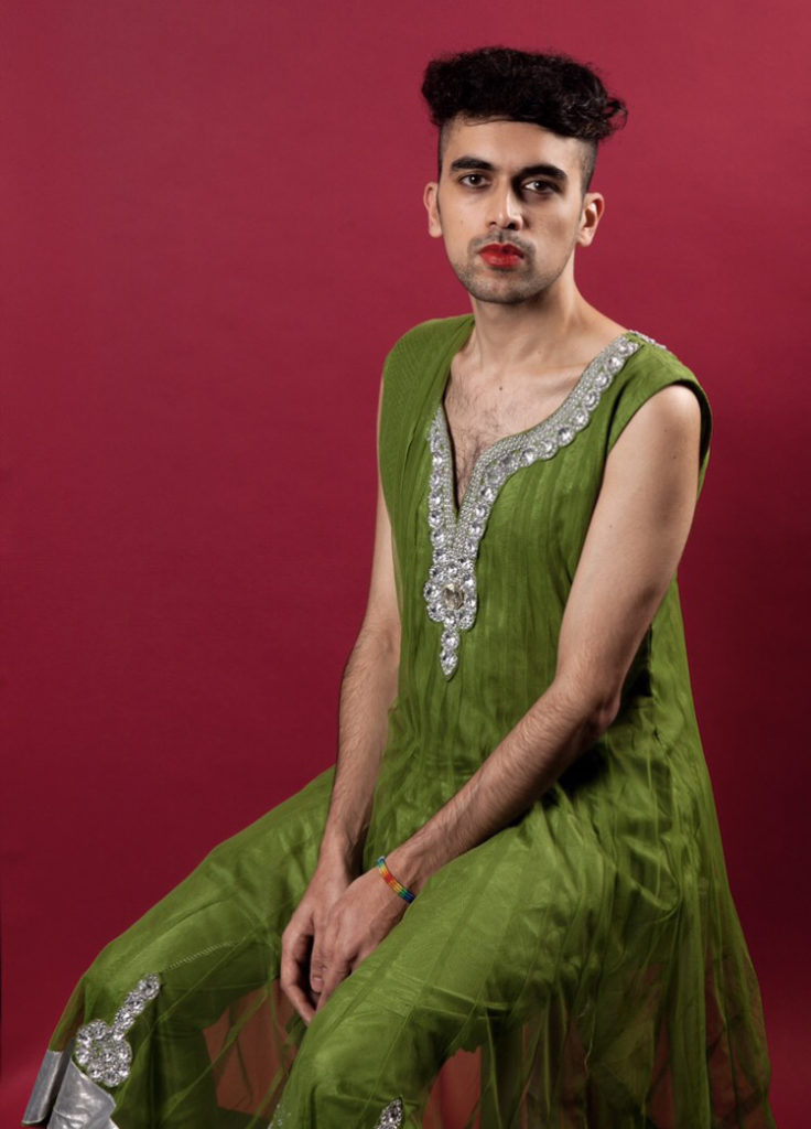 This is a photo of Vijay taken by Holly Revell. Vijay is a brown person with black hair. He is wearing a leafy green short sleeve saree with jewell detailing. He has red lipstick on and black eyeliner. The background is red. He is sitting down on a chair, his legs slightly apart and his hands are placed together on his lap.