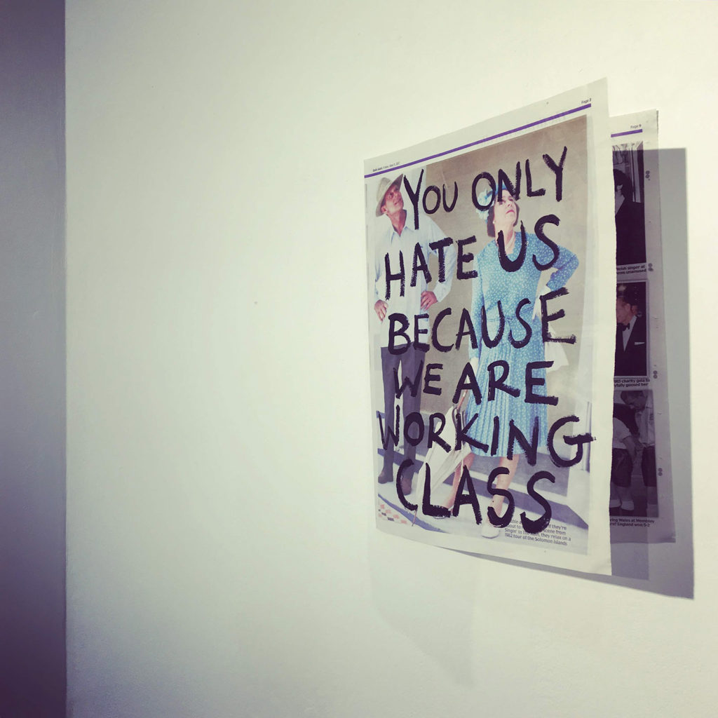 A leaf of a tabloid newspaper is stuck on a white wall so that the images on the inner page are visible at an angle, and causing a shadow against the wall. On the front page of the newspaper is a full-size photograph of a man and a woman. Written in black paint over the photograph are the words ‘YOU ONLY HATE US BECAUSE WE ARE WORKING CLASS’