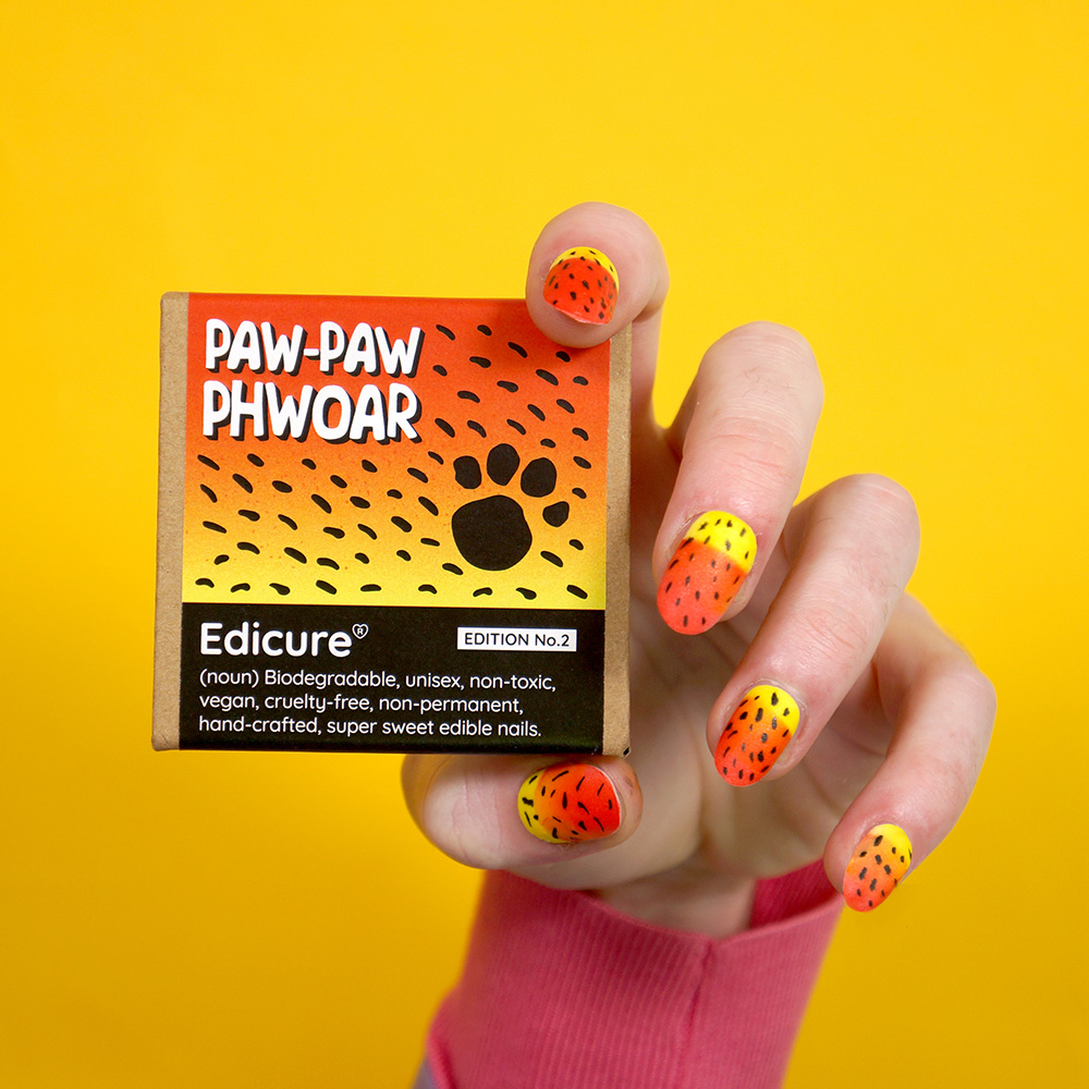 Against a bright yellow background, a white-skinned hand with yellow and orange animal print nails holds a small cardboard decorated with similar colours and a black cartoon paw print. The box says Paw-Paw Phwoar in large white letters at the top. Smaller text on the lower box reads: Edicure (noun): Biodegradable, unisex, non-toxic, vegan, cruelty-free, non-permanent, hand-crafted, super sweet edible nails.