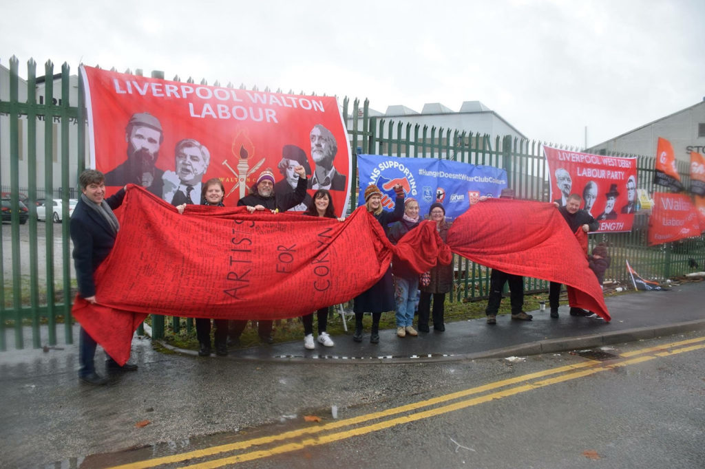 A group of smiling men and women are stood at the side of the road on rainy day. Between them they are holding a long red banner on which “Artist for Corbyn” is written in black, and behind them assorted political and campaigning banners hang from a metal fence